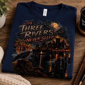 May include: A navy blue t-shirt featuring a graphic print of a city skyline at night with a full moon, a bridge, and a black raven perched in the foreground. The text reads THE THREE RIVERS NEVER SLEEP in a stylized, weathered orange and white font.