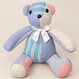 Könnte beinhalten: Ein Stoffteddybär aus Patchwork-Stoffbahnen in Blau-, Beige- und Rosatönen. Der Bär weist gestreifte und einfarbige Muster, schwarze Knopfaugen, eine schwarze Nase und eine weiße Satinschleife um den Hals auf.