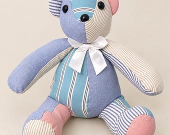 May include: A stuffed teddy bear constructed from patchwork fabric panels in shades of blue, beige, and pink. The bear features striped and solid patterns, black button eyes, a black nose, and a white satin ribbon tied around its neck.