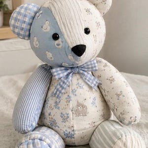 Può includere: Un orsacchiotto di pezza realizzato con vari ritagli di tessuto nelle tonalità del blu e del bianco. L'orso presenta motivi che includono quadretti vichy, righe e stampe floreali con piccoli animali. Un fiocco a quadretti blu e bianco è legato al collo.