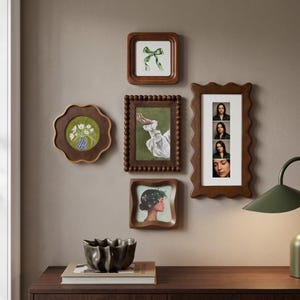 Op de afbeelding: Een galerijwand met vijf houten lijsten in verschillende vormen en maten. De lijsten bevatten kunstwerken, waaronder een groen lint, een hand in een witte mouw, een portret en een verticale strook met vier foto's van een persoon.
