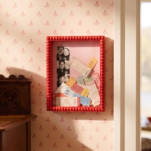 May include: A rectangular red shadow box frame with a beaded border containing a vertical strip of three black and white photographs and a collection of various colourful paper tickets and stubs arranged against a light pink backing.