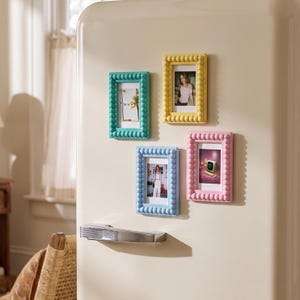 Puede incluir: Cuatro marcos de fotos magnéticos rectangulares con un diseño de borde de cuentas, cada uno en un color pastel diferente: verde azulado, amarillo, azul claro y rosa. Estos marcos están sujetos a la puerta de un refrigerador color crema.