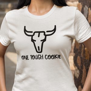 May include: White t-shirt featuring a black bull skull graphic and the text "ONE TOUGH COOKIE". The shirt has short sleeves and a crew neck. The design is centered on the chest.