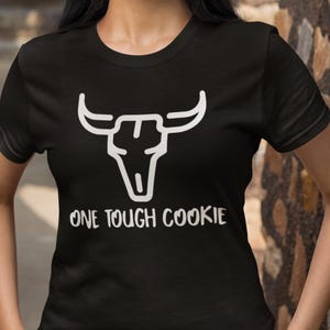 May include: Black t-shirt featuring a white graphic of a bull skull and the text "ONE TOUGH COOKIE". The shirt is a crew neck style and appears to be made of a soft material.