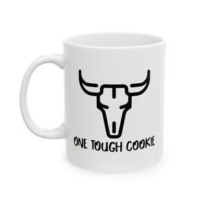 May include: White ceramic mug with a black graphic of a bull skull and the text "ONE TOUGH COOKIE". The mug has a curved handle and a cylindrical shape, suitable for hot or cold beverages.