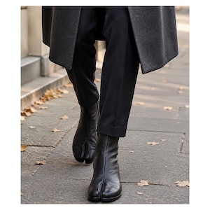 May include: A pair of black leather ankle boots featuring a distinct split-toe design, worn with black trousers and a dark gray coat. The boots have a smooth finish, a central seam running up the front, and a low heel, providing a sleek appearance.