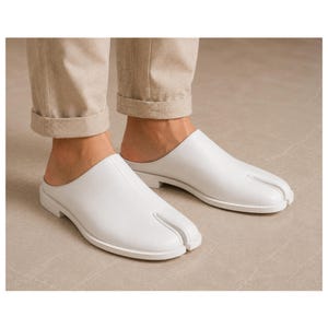 May include: A pair of white leather slip-on shoes featuring a distinct split-toe design. The shoes have a smooth finish, a low heel, and a clean, minimalist silhouette that covers the top of the foot while leaving the heel area exposed.