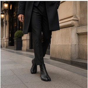 May include: A pair of black leather ankle boots featuring a distinct split-toe design, worn with tailored black trousers and a long black overcoat. The boots have a smooth finish, a side zipper closure, and a low block heel, providing a sleek appearance.