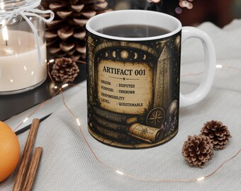 May include: A white ceramic mug featuring a dark, intricate design with the text ARTIFACT 001, ORIGIN: DISPUTED, PURPOSE: UNKNOWN, RESPONSIBILITY LEVEL: QUESTIONABLE, and the phrases ANCIENT MYSTERIES, UNEXPLAINED, and STILL LEARNING printed on the side.