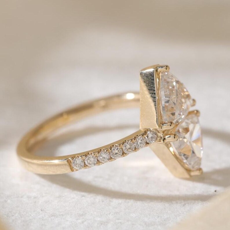 May include: A gold ring featuring two pear-shaped diamonds. The band is adorned with small, round diamonds. The ring is set against a soft, neutral background, highlighting the sparkle of the gemstones.
