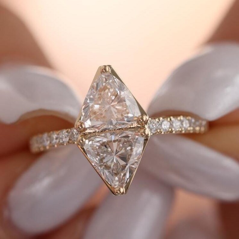 May include: A gold ring featuring two large, clear, triangular-cut gemstones stacked on top of each other. The band is also gold and encrusted with small, clear gemstones. The ring is held up against a soft, blurred background.