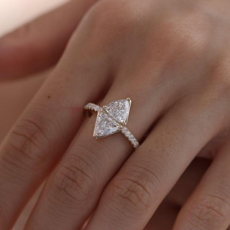 May include: A gold ring featuring a large, kite-shaped diamond as the centerpiece. The band is also adorned with smaller, sparkling diamonds. The ring is set against a blurred background, highlighting the jewelry.