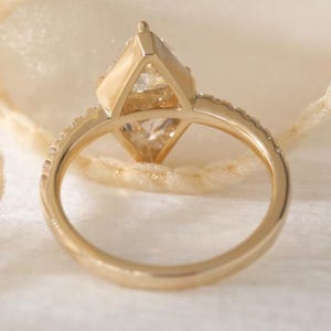 May include: A gold ring with a large, clear, kite-shaped gemstone as the centerpiece. The band is also gold and has small, clear stones set along the top half. The ring is set against a soft, off-white background.