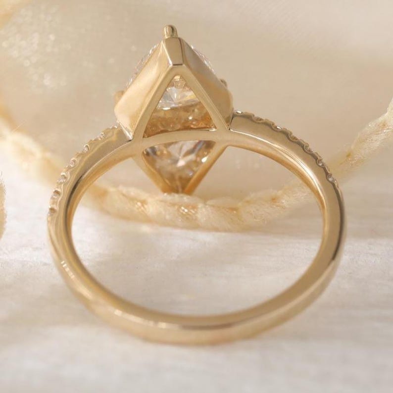 May include: A gold ring with a large, clear, kite-shaped gemstone as the centerpiece. The band is also gold and has small, clear stones set along the top half. The ring is set against a soft, off-white background.