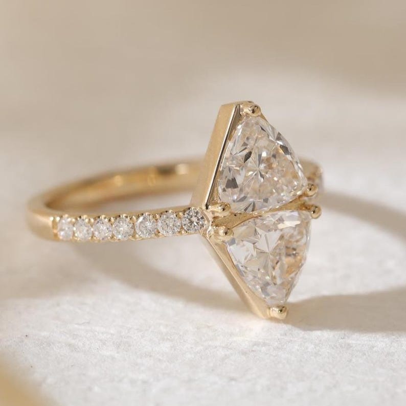 May include: A gold ring featuring two large, clear, triangular-cut gemstones. The band is gold and has a row of small, round, clear gemstones. The ring is set against a soft, neutral background.