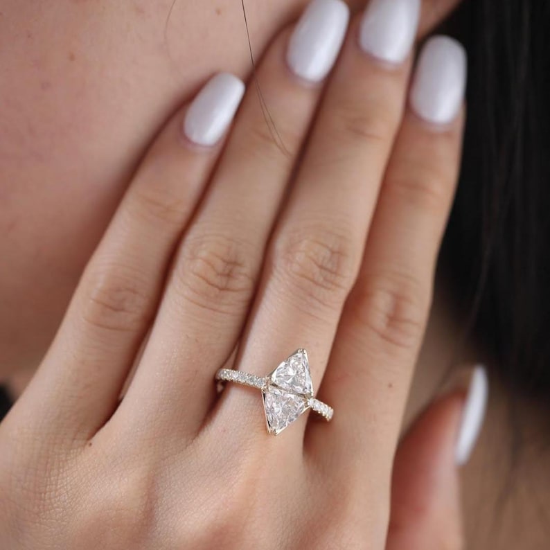 May include: A gold ring featuring a large, triangular-cut diamond as the centerpiece. The band is adorned with smaller, pav&eacute;-set diamonds, adding sparkle. The ring is shown on a hand with white nail polish.