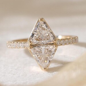 May include: A gold ring featuring two large, triangular-cut diamonds set side-by-side. The band is adorned with smaller, round diamonds. The ring is set on a white surface, showcasing its sparkle and intricate design.