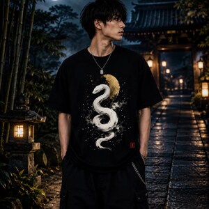 Può includere: Una t-shirt nera a maniche corte con una stampa grafica di un serpente bianco avvolto su una luna dorata e un cielo stellato. Il design include un piccolo emblema quadrato rosso sul lato inferiore destro. La t-shirt è abbinata a pantaloni cargo neri.