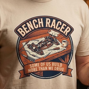 May include: A beige t-shirt featuring a circular graphic with the text "BENCH RACER" and "SOME OF US BUILD MORE THAN WE DRIVE." The illustration depicts a remote-controlled car on a workbench with tools and a mug, rendered in orange and blue.