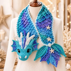 Könnte beinhalten: Ein gehäkelter Schal mit Drachenkopf und Meerjungfrauenschwanz in Blau-, Türkis- und Violetttönen. Der Schal hat eine schuppenartige Textur, weiße Akzente am Drachenkopf und drei kleine Seestern-Applikationen am Schwanzende des Schals.