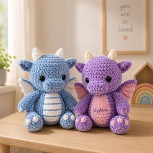 Könnte beinhalten: Zwei gehäkelte Drachen-Stofftiere. Der linke Drache ist blau mit weißen Hörnern, Flügeln und einem gestreiften Bauch. Der rechte Drache ist lila mit cremefarbenen Hörnern, rosa Flügeln und einem rosa Bauch mit dem gestickten Namen Wyshine.