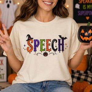 Può includere: T-shirt color crema con la scritta "SPEECH" in lettere colorate a tema Halloween. Il design include un cappello da strega, un pipistrello e una zucca. Una tazza nera a forma di zucca è tenuta in mano.