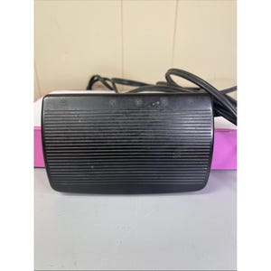 Könnte beinhalten: Schwarzes Singer-Nähmaschinen-Fußpedal mit geriffelter Oberfläche. Das Wort "SINGER" ist oben aufgedruckt. Ein schwarzes Netzkabel erstreckt sich von dem Pedal.