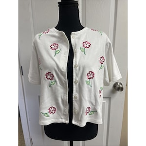 May include: White short-sleeved cardigan with embroidered flowers in shades of pink and green. The cardigan has a button-up front and a round neckline. The floral design is repeated across the front and shoulders of the garment.