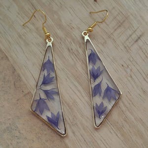 Könnte beinhalten: Ein Paar Ohrhänger mit dreieckigen, goldfarbenen Metallrahmen, die mit klarem Harz gefüllt sind. Jeder Ohrring enthält mehrere kleine, gepresste violette Blüten, die im Harz eingebettet sind und an goldfarbenen Metall-Ohrhaken befestigt sind.