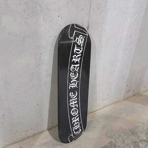 May include: Black skateboard with the text "CHROME HEARTS" in white script. The skateboard is leaning against a gray wall. The skateboard is a long, narrow board with rounded ends.
