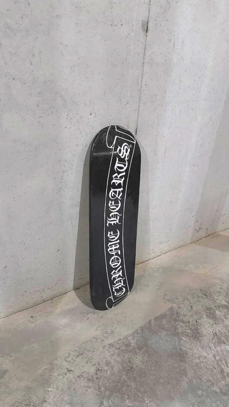 May include: Black skateboard with the text "CHROME HEARTS" in white script. The skateboard is leaning against a gray wall. The skateboard is a long, narrow board with rounded ends.