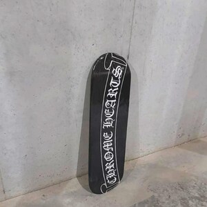 May include: Black skateboard with the text "CHROME HEARTS" in white script. The skateboard is leaning against a gray concrete wall. The board has a glossy finish.