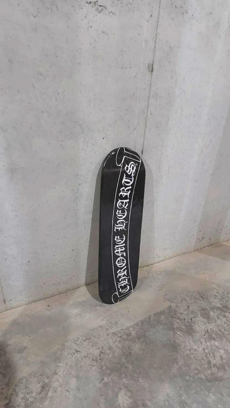 May include: Black skateboard with the text "CHROME HEARTS" in white script. The skateboard is leaning against a gray concrete wall. The board has a glossy finish.