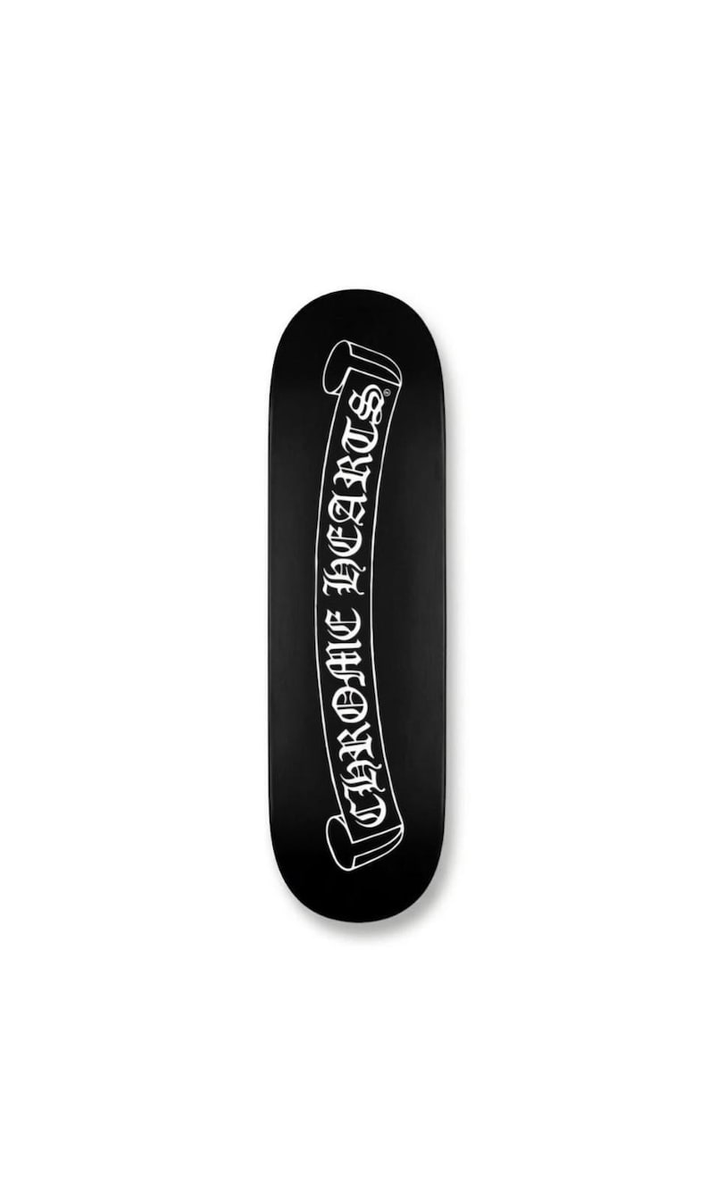May include: Black skateboard with a white banner design. The banner features the text "CHROME HEARTS" in a gothic font. The skateboard has a rounded shape and is set against a white background.