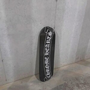 May include: Black skateboard with the text "CHROME HEARTS" in white script. The skateboard is positioned vertically against a gray concrete wall. The design is a classic, bold statement.