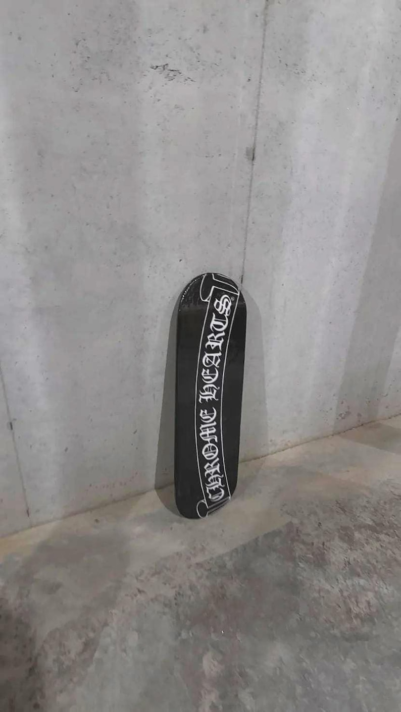 May include: Black skateboard with the text "CHROME HEARTS" in white script. The skateboard is positioned vertically against a gray concrete wall. The design is a classic, bold statement.