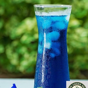 Könnte beinhalten: Ein hohes, klares Glas gefüllt mit einer leuchtend blauen Flüssigkeit und Eiswürfeln, mit Kondenswasser an der Außenseite. Neben dem Glas liegen zwei blaue Schmetterlingserbsenblüten. Ein kreisförmiges Logo mit dem Text CEYLON GREEN HERBALS ist sichtbar.