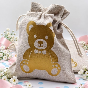 Puede incluir: Una bolsa con cordón de color beige hecha de tela texturizada que presenta un gráfico dorado metálico de un oso de peluche sentado con pajarita. La bolsa tiene cordones de tela beige a juego en la parte superior para cerrar la abertura.