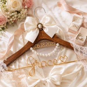 May include: A wooden wedding dress hanger with a cream-colored bow and pearl accents. The hanger features the name "Grace" in gold wire and the date "05.18.2027". Two ring boxes are visible in the background.