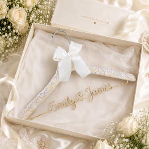 May include: A clothes hanger covered in cream fabric with a silver floral pattern, featuring a white satin ribbon bow at the top and gold wire lettering that reads Emily & James, presented inside a cream gift box with the text FOR YOUR SPECIAL DAY.