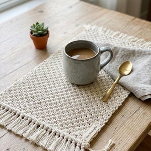 May include: A cream-coloured macrame placemat with a fringed edge, a grey ceramic mug filled with coffee, and a gold spoon on a wooden table. A small succulent in a terracotta pot is also visible. The placemat has a tag that says "Shop Handcrafted".