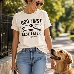 May include: Cream-colored t-shirt with the text "DOG FIRST. Everything ELSE LATER." in black. The shirt features a paw print and a heart symbol. The person is wearing blue jeans and sunglasses, walking a golden retriever dog.