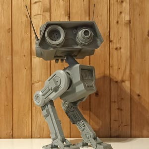 Op de afbeelding: Een grijze robotfiguur met een rechthoekig hoofd, twee cirkelvormige, oogachtige lenzen en twee dunne antennes. De robot heeft een centrale romp verbonden met twee gearticuleerde mechanische benen met zichtbare gewrichten en voeten.