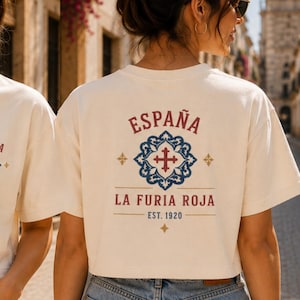 May include: A cream-colored short-sleeve t-shirt featuring a small graphic on the front left chest and a larger matching graphic on the back. The design includes the text ESPAÑA, LA FURIA ROJA, and EST. 1920 with a blue and red ornate cross emblem.