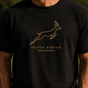 A man wearing a black t-shirt with a gold outline of a jumping springbok and the words "SOUTH AFRICA AMABOKOBOKO" below it, standing on a rugby field.