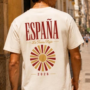 Op de afbeelding: Een off-white t-shirt met korte mouwen met de rode tekst ESPAÑA, de cursieve tekst La Furia Roja, een cirkelvormige zonnestraalgrafiek in rood en goud, en het jaartal 2026 op de achterkant. Het shirt heeft een ronde hals en een relaxte pasvorm.