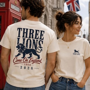 Könnte beinhalten: Cremefarbene T-Shirts mit einer dunkelblauen Löwengrafik. Auf der Rückseite stehen 'THREE LIONS', 'Come On England', '1966' und '2026'. Vorne befindet sich ein kleines Löwensymbol und der Schriftzug 'THREE LIONS' auf der linken Brust.