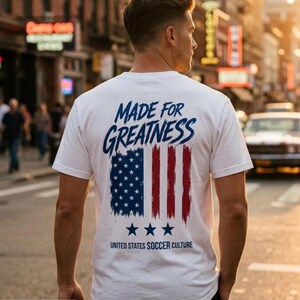 以下が含まれることがあります： 青い文字でMADE FOR GREATNESSと書かれ、赤と青の筆致のストライプが入ったアメリカ国旗のグラフィック、3つの青い星、そして下にUNITED STATES SOCCER CULTUREという文字がプリントされた白い半袖Tシャツ。