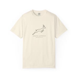 A cream-colored t-shirt with a black outline of a jumping springbok and the words "SOUTH AFRICA AMABOKOBOKO" below it, laid flat on a white background.
